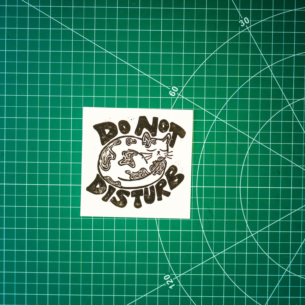 A square linocut design in black ink of a sleeping cat with the words "do not disturb" around it. It's displayed on a green cutting mat with grid lines.