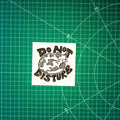 A square linocut design in black ink of a sleeping cat with the words "do not disturb" around it. It's displayed on a green cutting mat with grid lines.