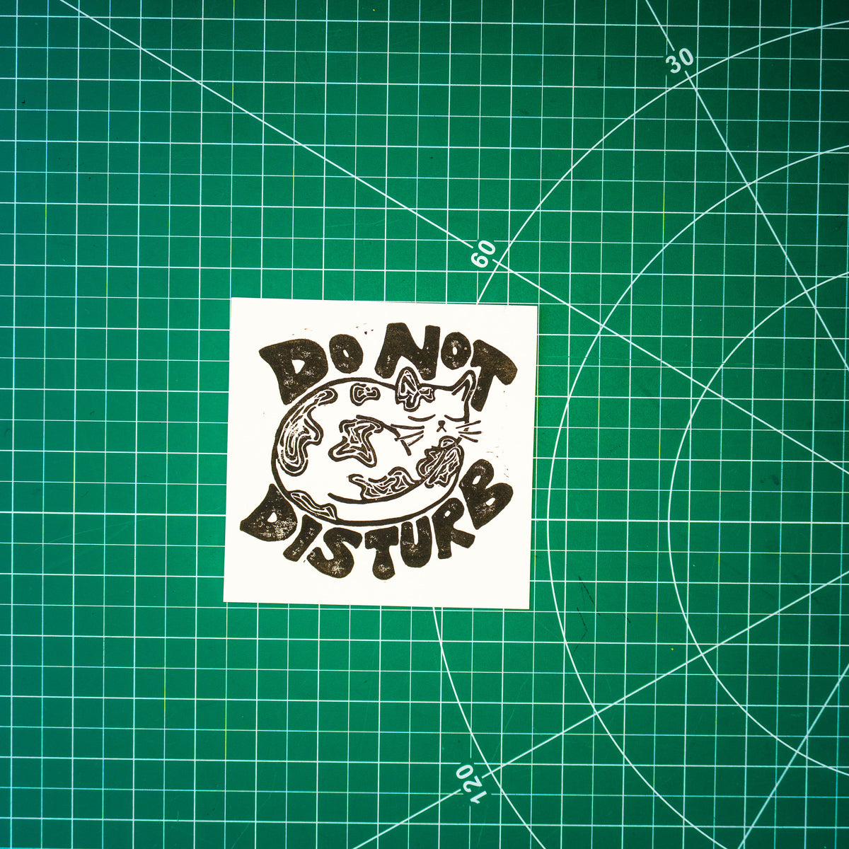 A square linocut design in black ink of a sleeping cat with the words "do not disturb" around it. It's displayed on a green cutting mat with grid lines.