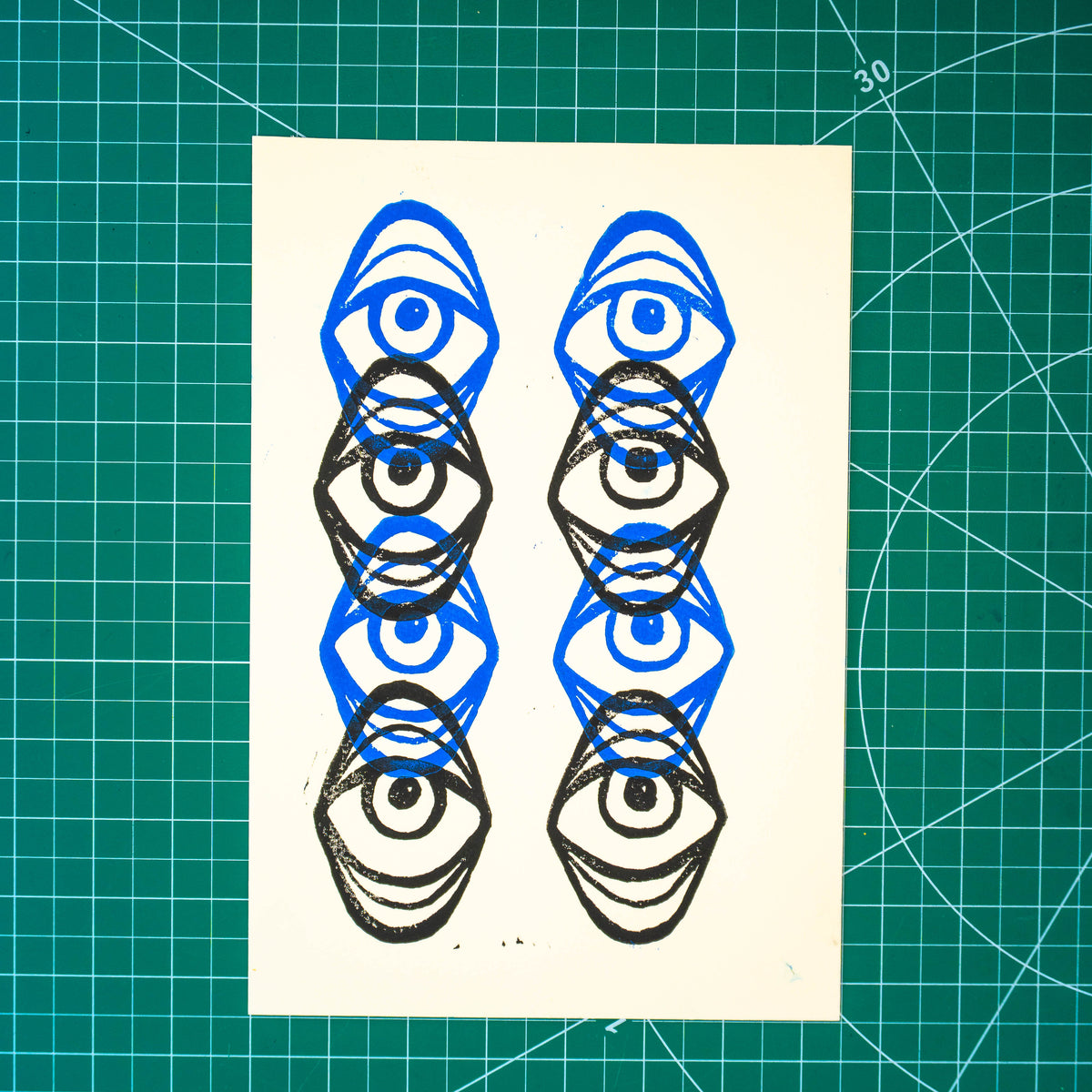 A square art print featuring six linocut eye designs in blue and black ink, displayed on a green cutting mat.