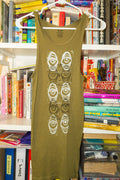 An olive green sleeveless tank top with a white and gray eye pattern down the chest, hanging on a white hanger in front of a bookshelf.