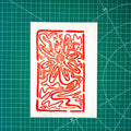 A red abstract floral or sunburst-style linocut/woodblock print on a white rectangular paper sheet, placed on a green gridded cutting mat.