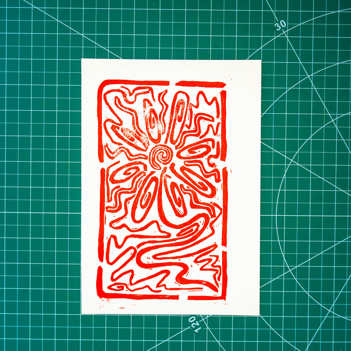 A red abstract floral or sunburst-style linocut/woodblock print on a white rectangular paper sheet, placed on a green gridded cutting mat.