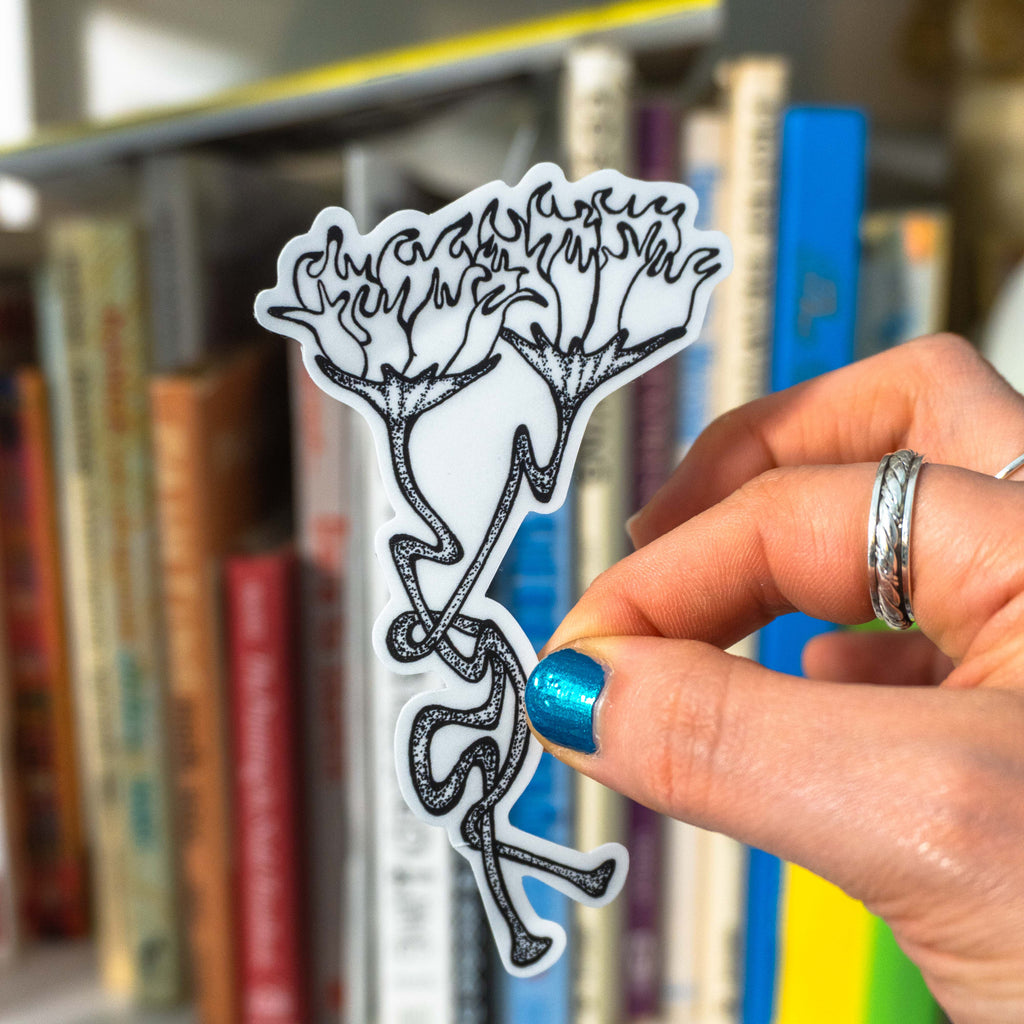 A hand with blue glitter nail polish holds a white die-cut sticker featuring a black line art illustration of two flowers with long stems, in front of a blurred bookshelf background.