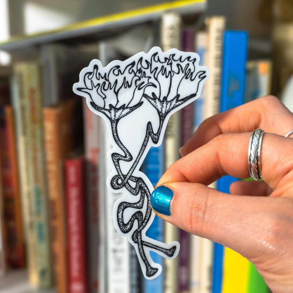 A hand with blue glitter nail polish holds a white die-cut sticker featuring a black line art illustration of two flowers with long stems, in front of a blurred bookshelf background.