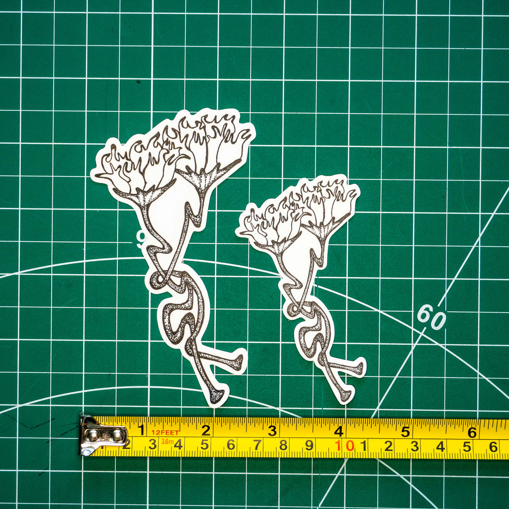Two differently-sized die-cut stickers featuring a grayscale tattoo-style design of two intertwining flowers placed on a green cutting mat with a yellow tape measure below for scale.