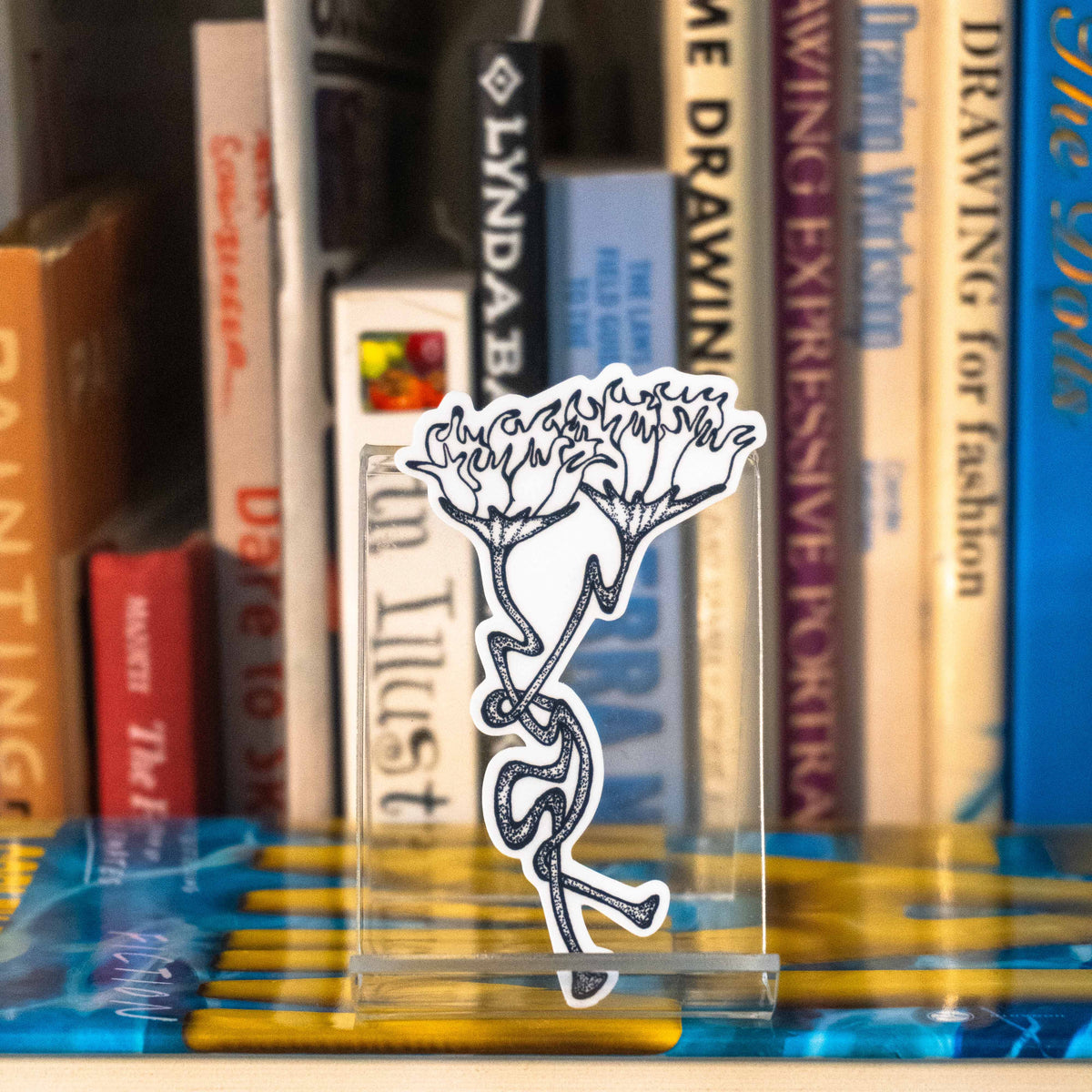A small die-cut sticker featuring a grayscale tattoo-style design of two intertwining flowers displayed on a clear acrylic stand in front of a bookshelf.