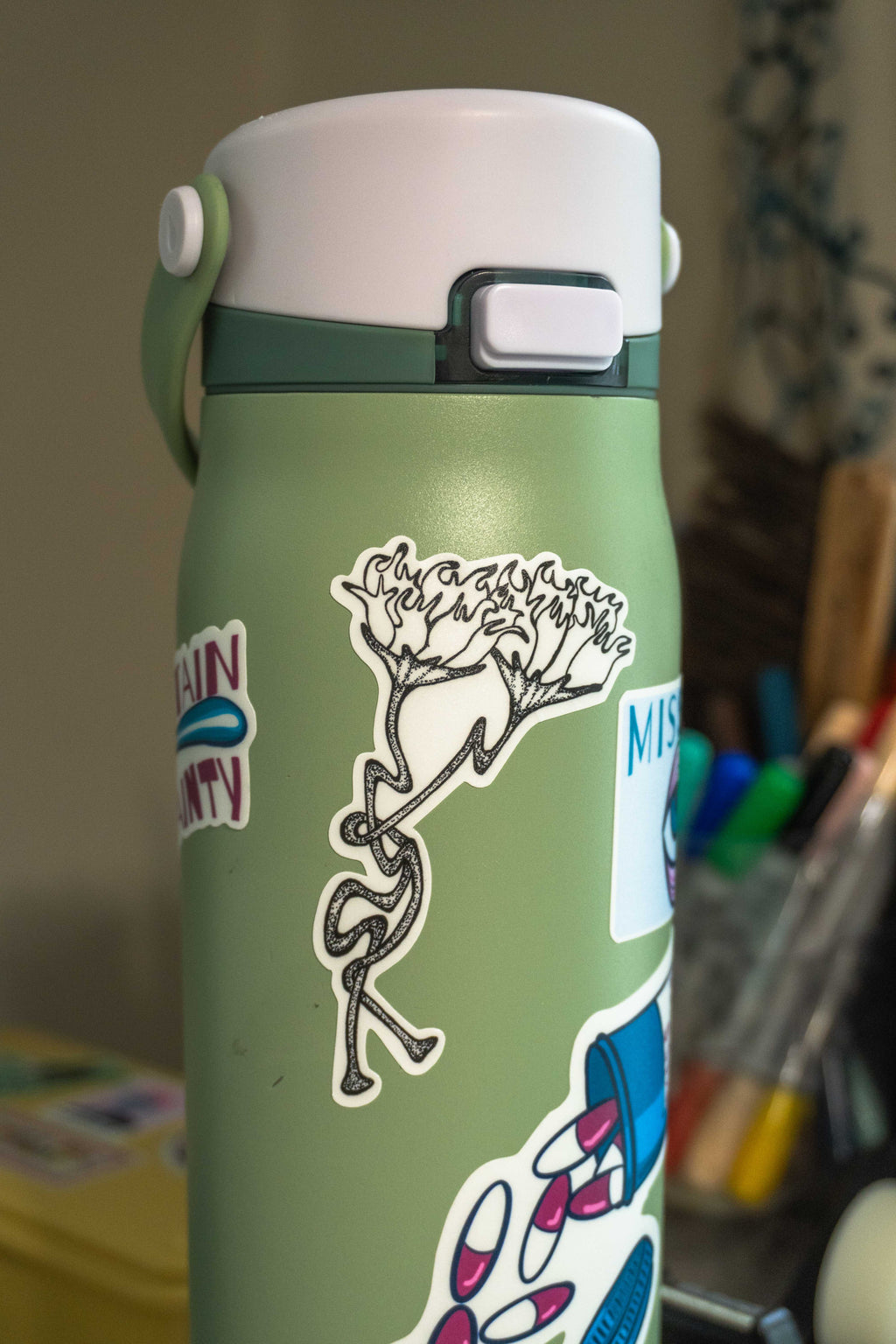 A light sage green insulated water bottle with a white lid, decorated with several stickers including the die-cut tattoo-style flower design in the center, shown in a blurred indoor setting.