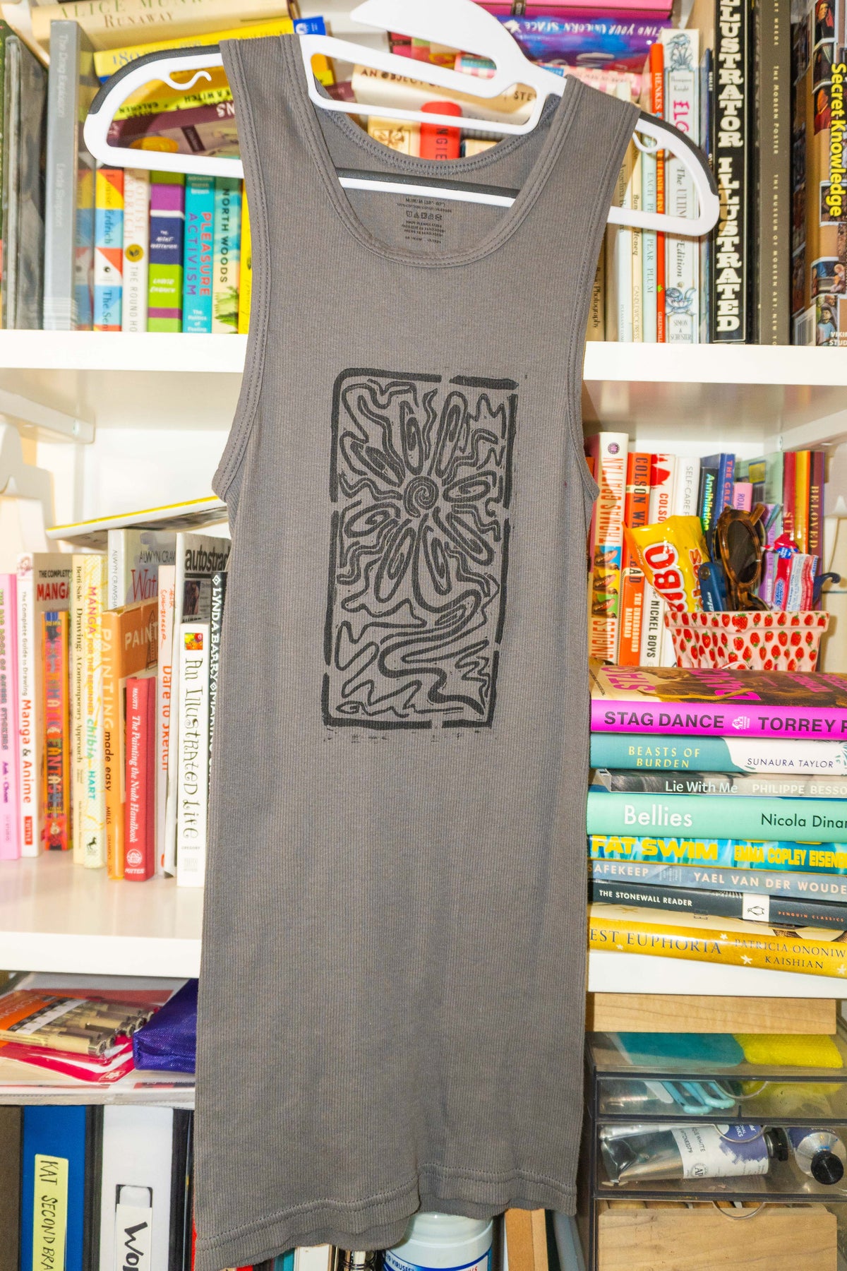A gray sleeveless tank top with a black linocut flower design hanging on a white hanger in front of a bookshelf filled with books.