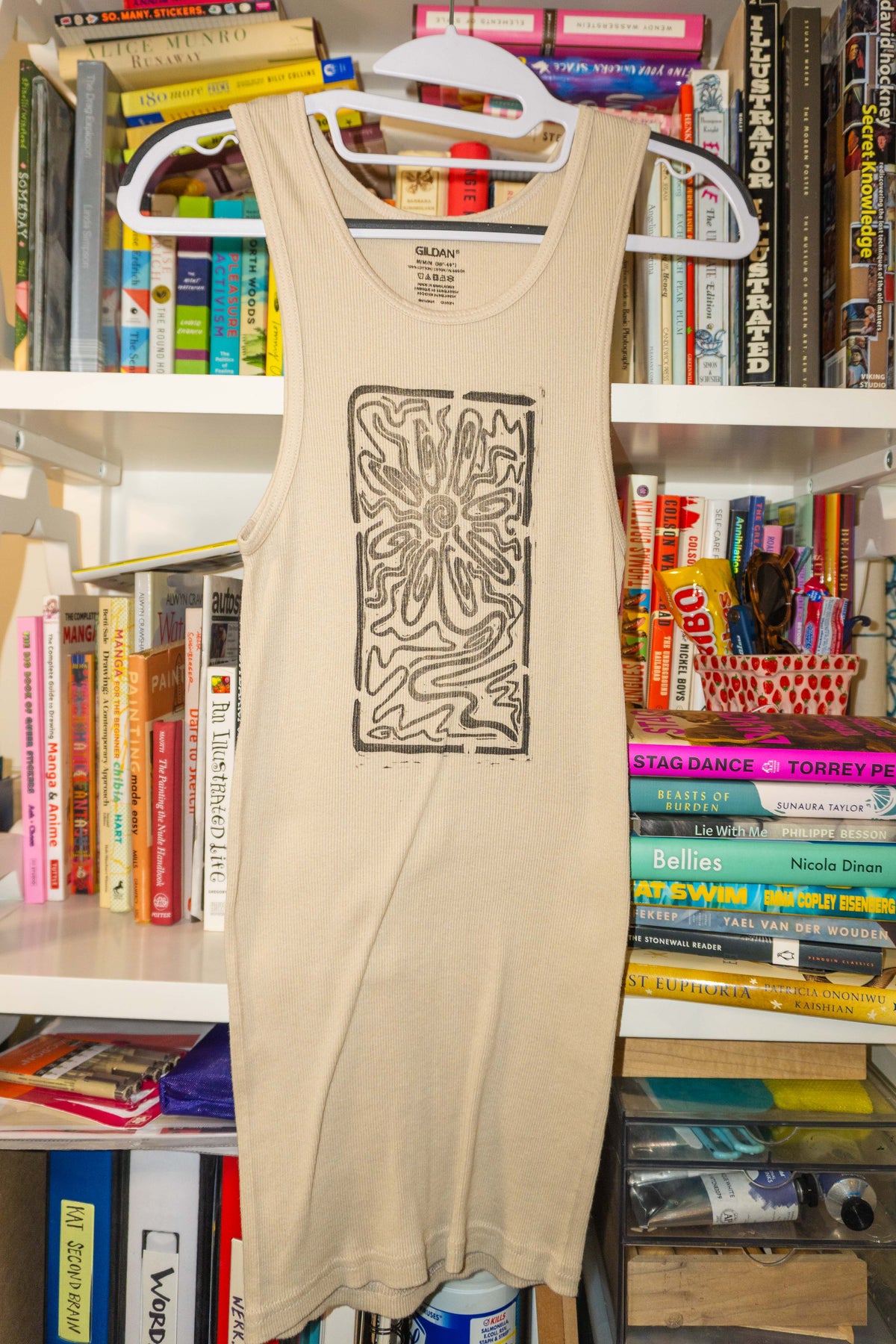 A beige tank top with a black rectangular linocut flower print hanging on a white hanger in front of a bookshelf filled with books.