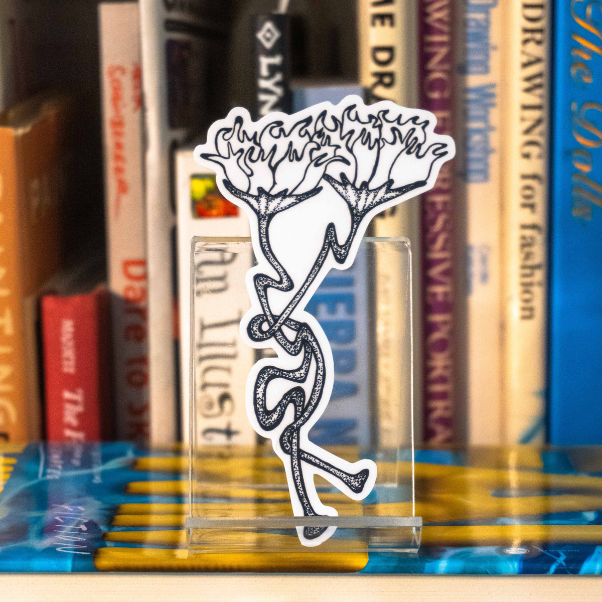 A large die-cut sticker featuring a grayscale tattoo-style design of two intertwining flowers displayed on a clear acrylic stand in front of a bookshelf.