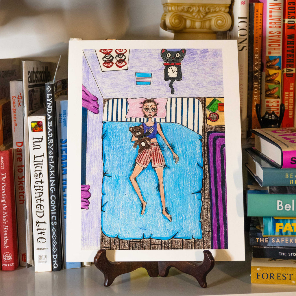 A colorful hand-drawn illustration on a white card, showing a girl lying on a blue bed with a teddy bear, displayed on a small wooden easel between books.
