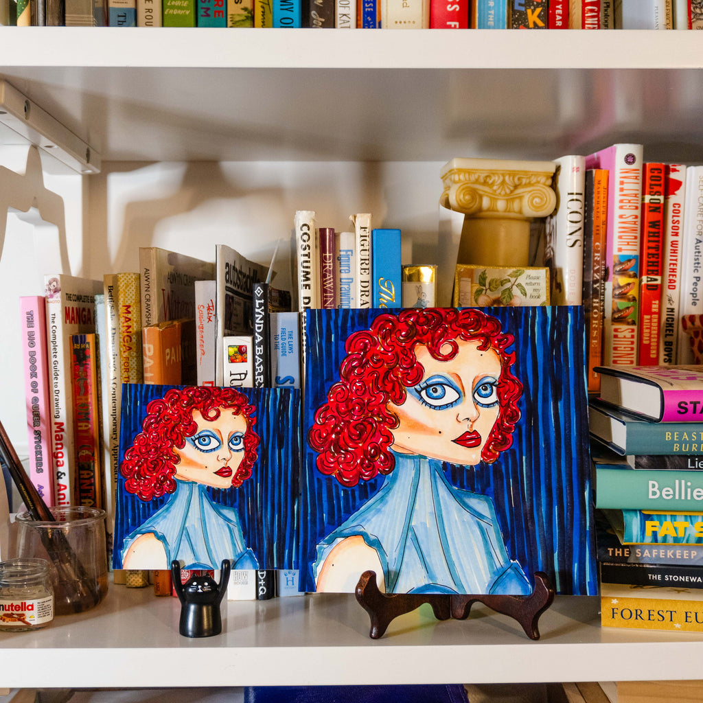 Two colorful paintings of a woman with bright red curly hair and blue eyes, displayed on a white bookshelf between books.