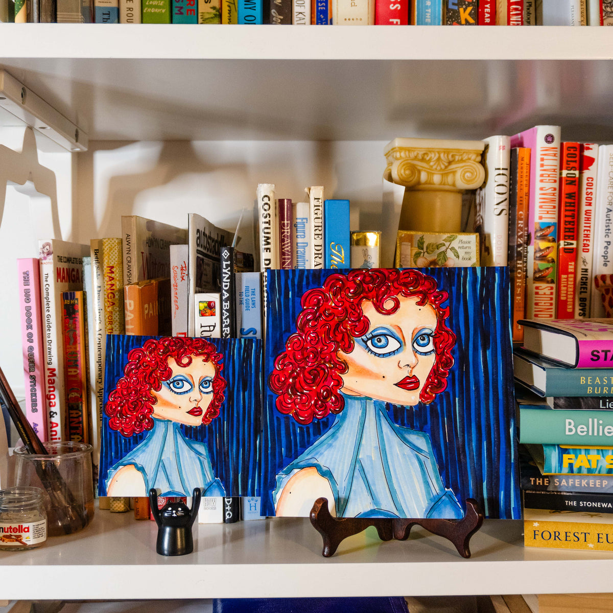 Two colorful paintings of a woman with bright red curly hair and blue eyes, displayed on a white bookshelf between books.