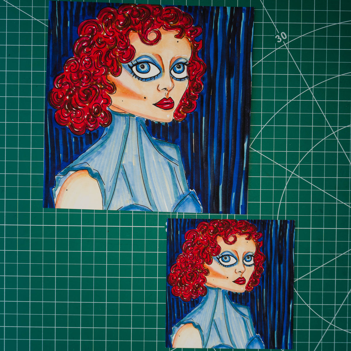 Two hand-drawn portraits of a woman with bright red curly hair, blue eyes, and a light blue dress, set against dark blue striped backgrounds. One larger portrait is positioned in the upper left, and a smaller version is in the lower right, both placed on a green cutting mat with grid lines.