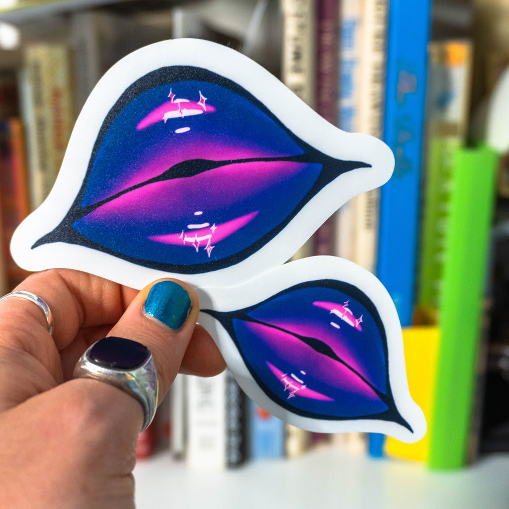 Hand with blue nail polish and silver rings holding both size variations of die-cut lips sticker in front of a bookshelf.