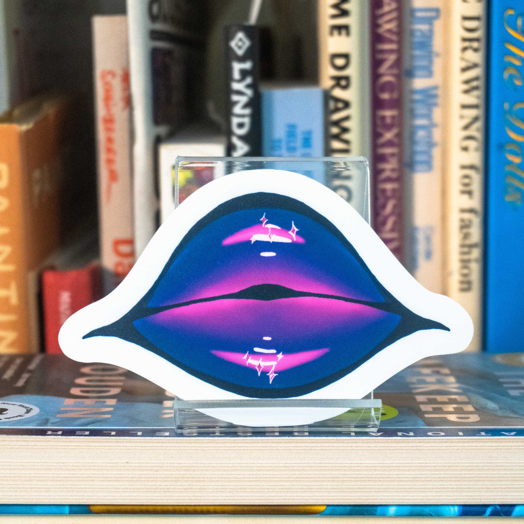 A large die-cut sticker featuring illustrated lips covered in purple and pink glittering lipstick displayed on a clear acrylic stand in front of a bookshelf.