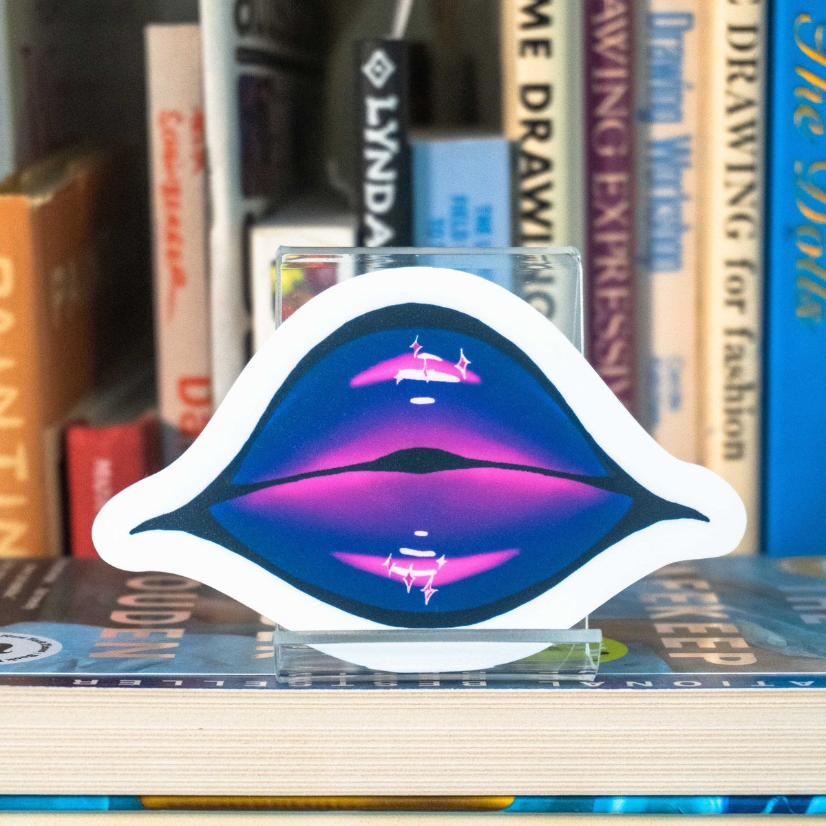 A large die-cut sticker featuring illustrated lips covered in purple and pink glittering lipstick displayed on a clear acrylic stand in front of a bookshelf.