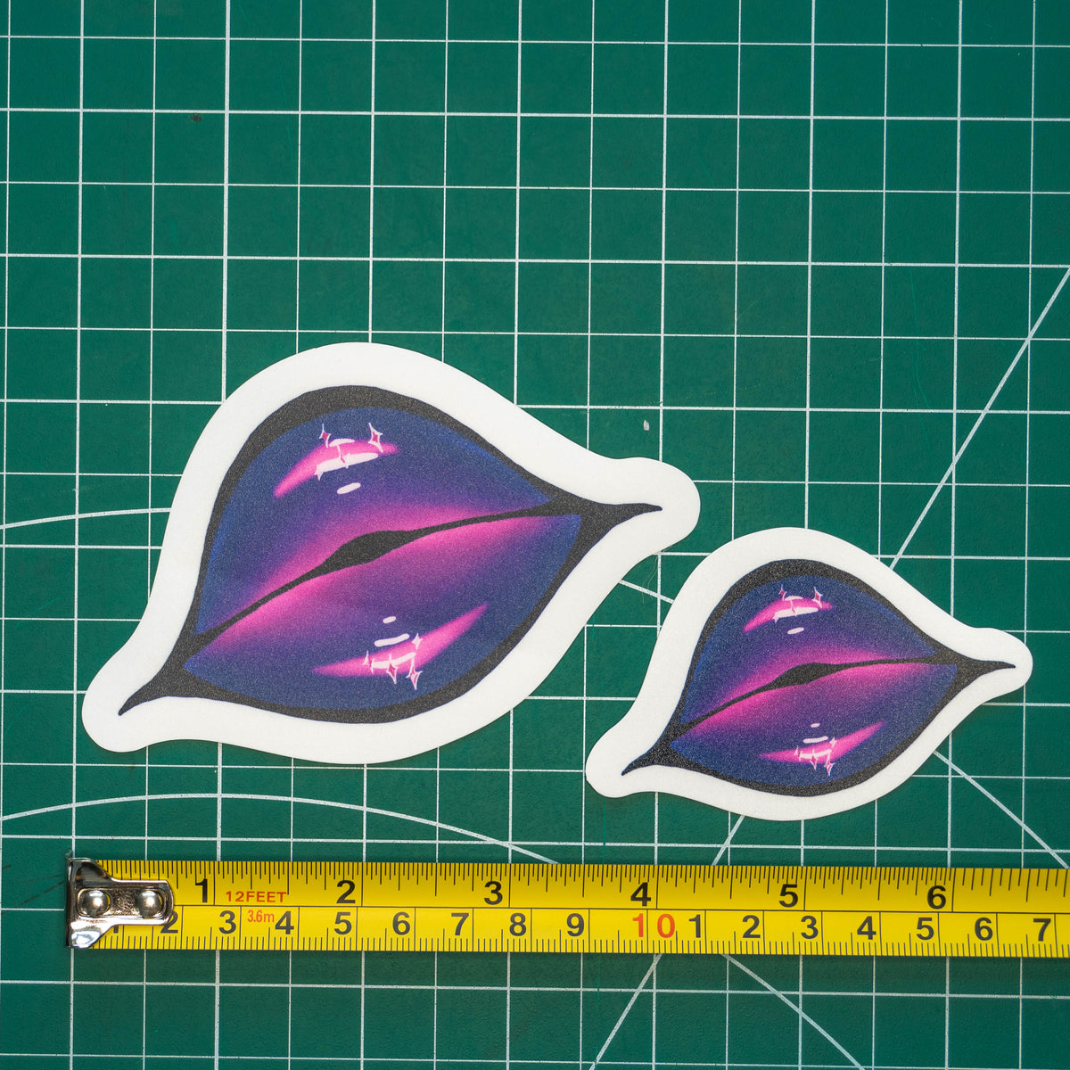 Two differently-sized die-cut stickers featuring illustrated lips covered in pink and purple glittering lipstick placed on a green cutting mat with a yellow tape measure below for scale.