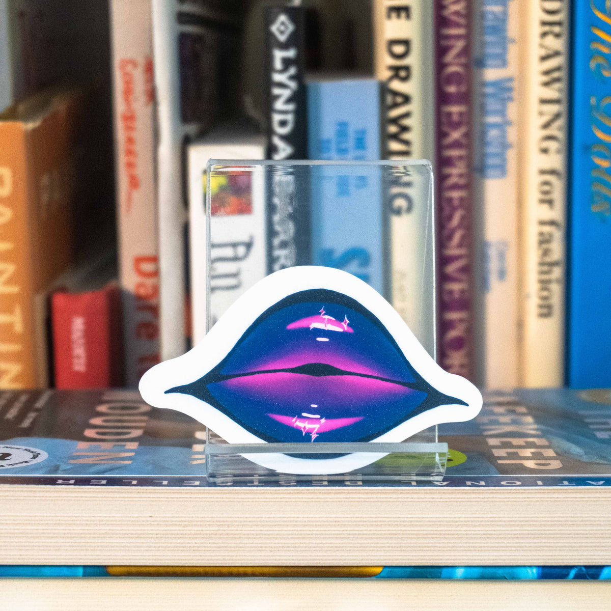 A small die-cut sticker featuring illustrated lips covered in purple and pink glittering lipstick displayed on a clear acrylic stand in front of a bookshelf.