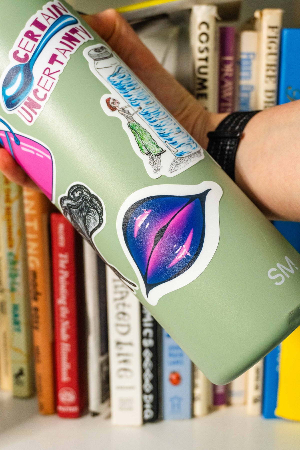 A hand holding a sage green insulated bottle decorated with colorful stickers and die-cut lips sticker in the center, set against a blurred background of book spines.