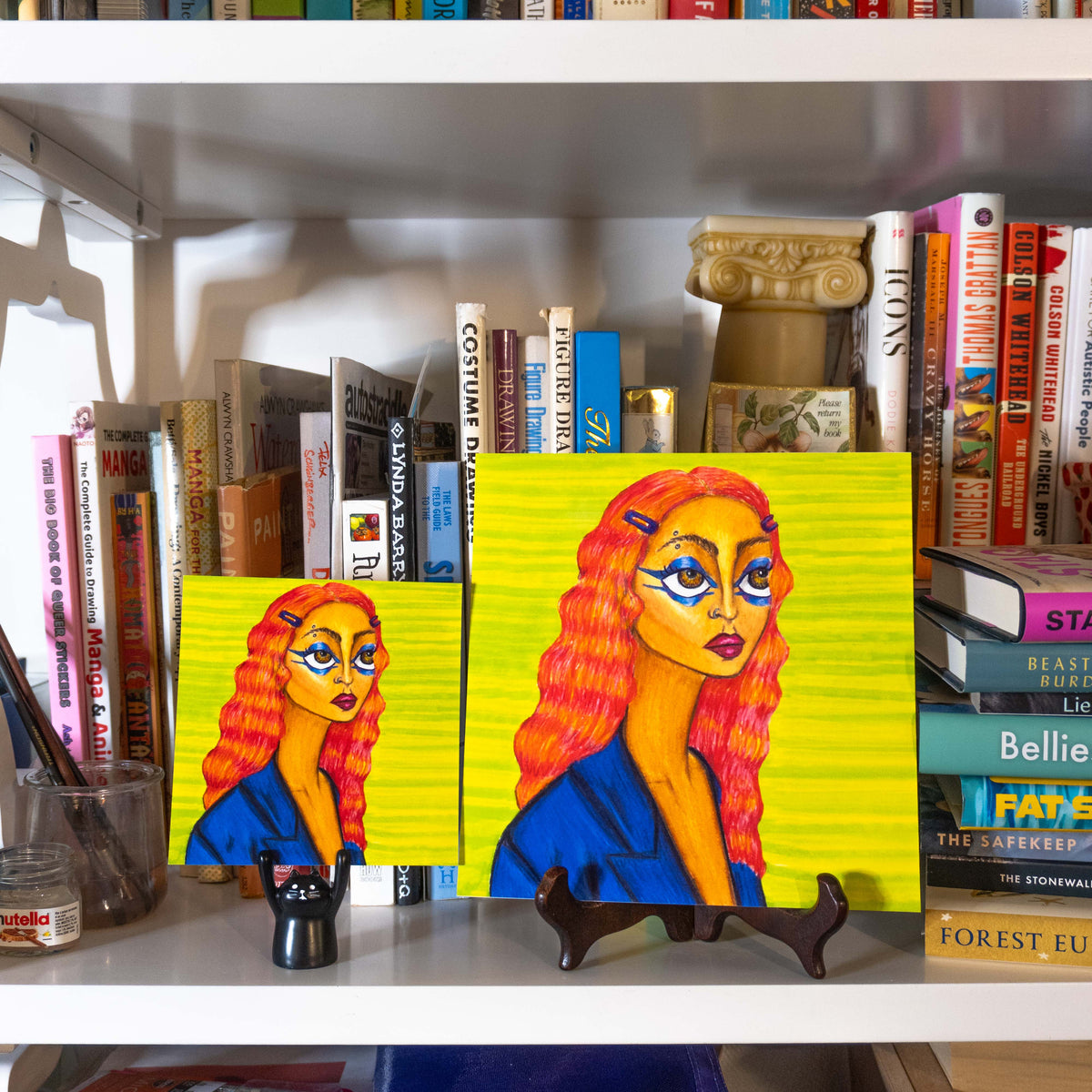 Two colorful portraits of a woman with bright yellow hair and blue clothing, displayed on a white bookshelf between books.