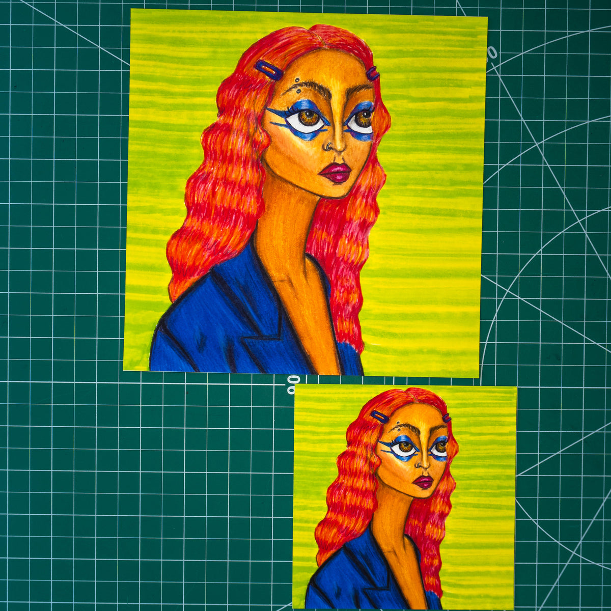 Two art prints in different sizes on a green cutting mat. The prints contain a colored pencil portrait of a woman’s bust in profile. She has caramel-colored skin, vivid blue eye makeup, a red lip, an eyebrow piercing, and striped red-and-orange hair pinned back with purple clips. 