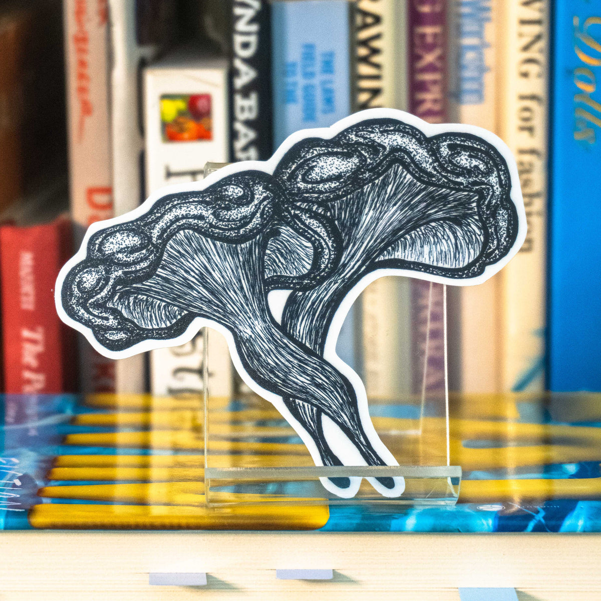 A large die-cut sticker featuring a grayscale tattoo-style illustration of a western jack-o-lantern mushroom displayed on a clear acrylic stand in front of a bookshelf.