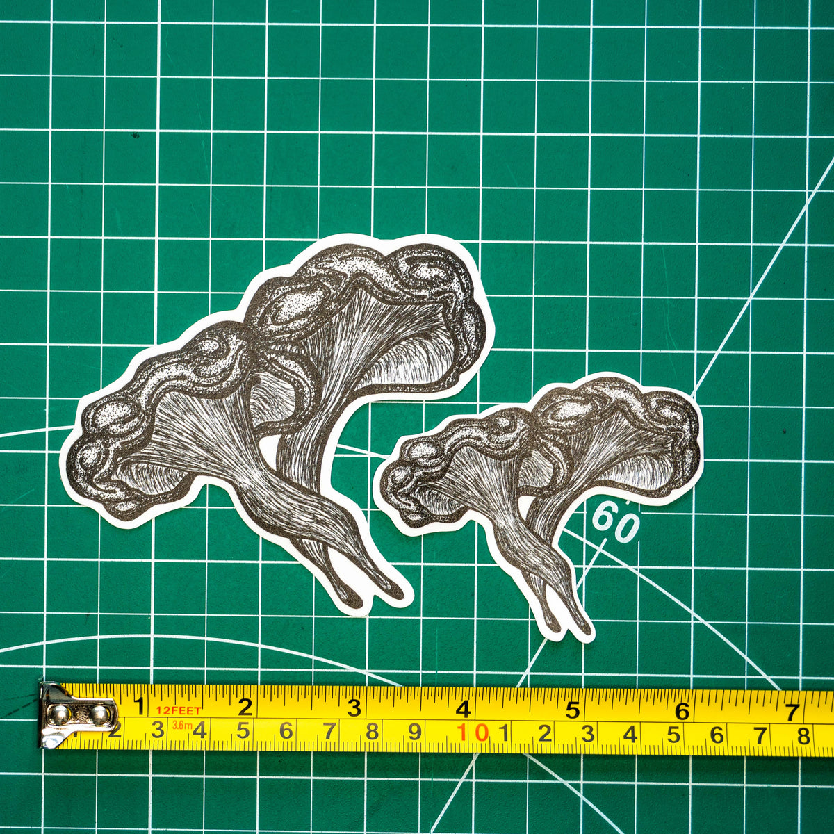 Two differently-sized die-cut stickers featuring a grayscale tattoo-style illustration of a western jack-o-lantern mushroom placed on a green cutting mat with a yellow tape measure below for scale.