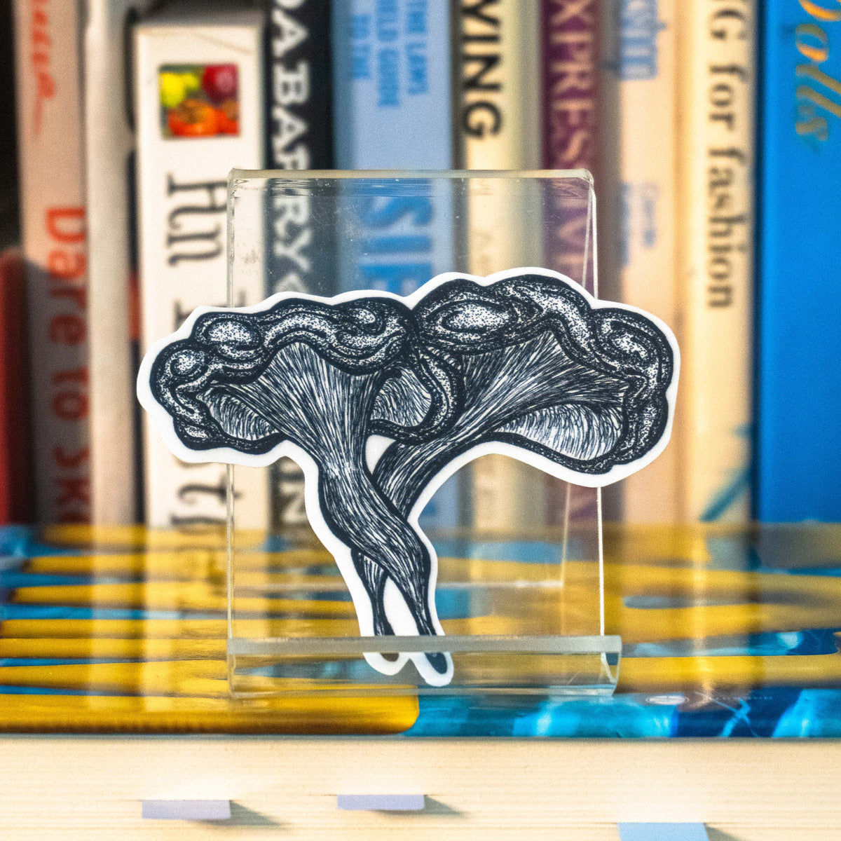 A small die-cut sticker featuring a grayscale tattoo-style illustration of a western jack-o-lantern mushroom displayed on a clear acrylic stand in front of a bookshelf.