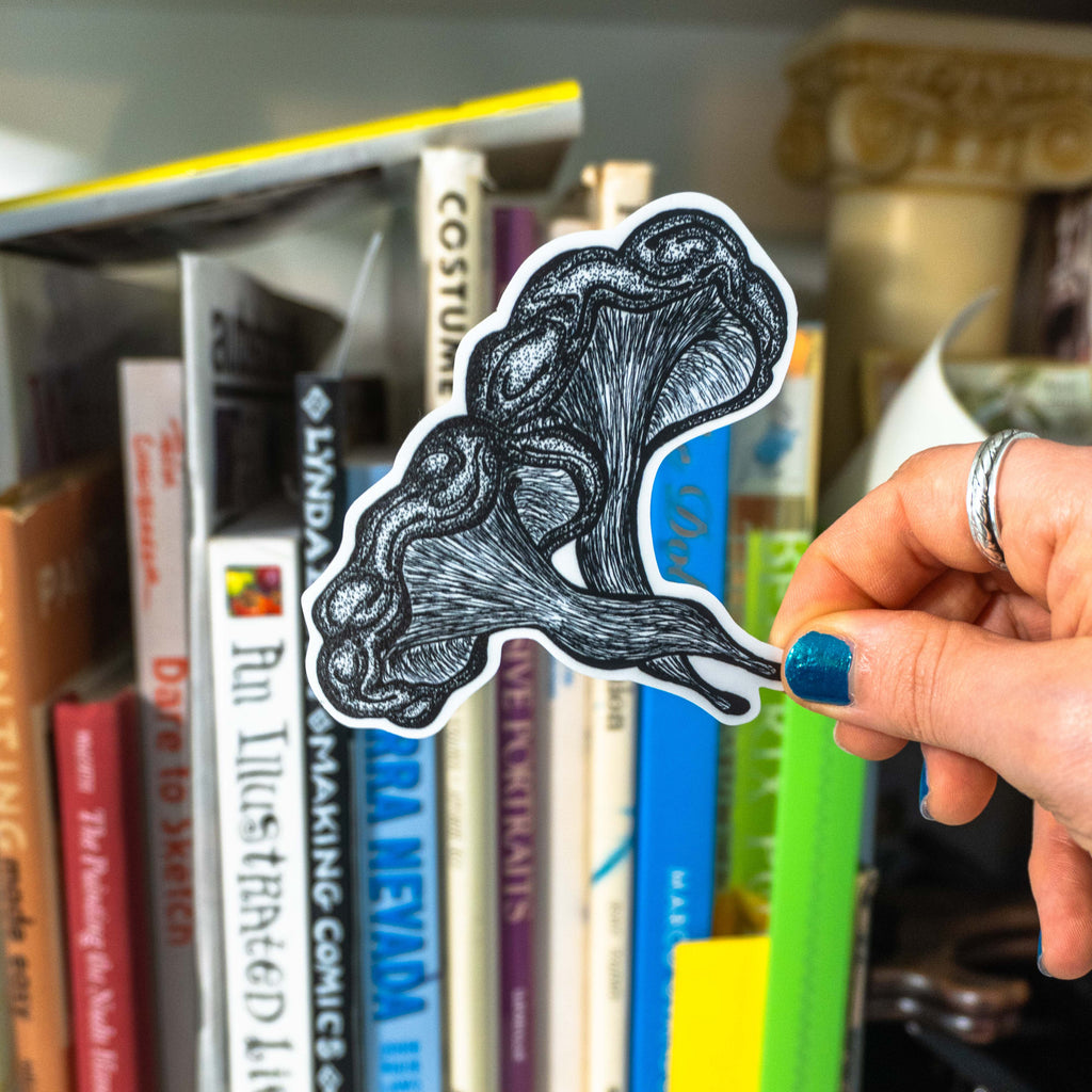 hand with blue nail polish holding the small die-cut mushroom sticker in front of a blurred bookshelf.