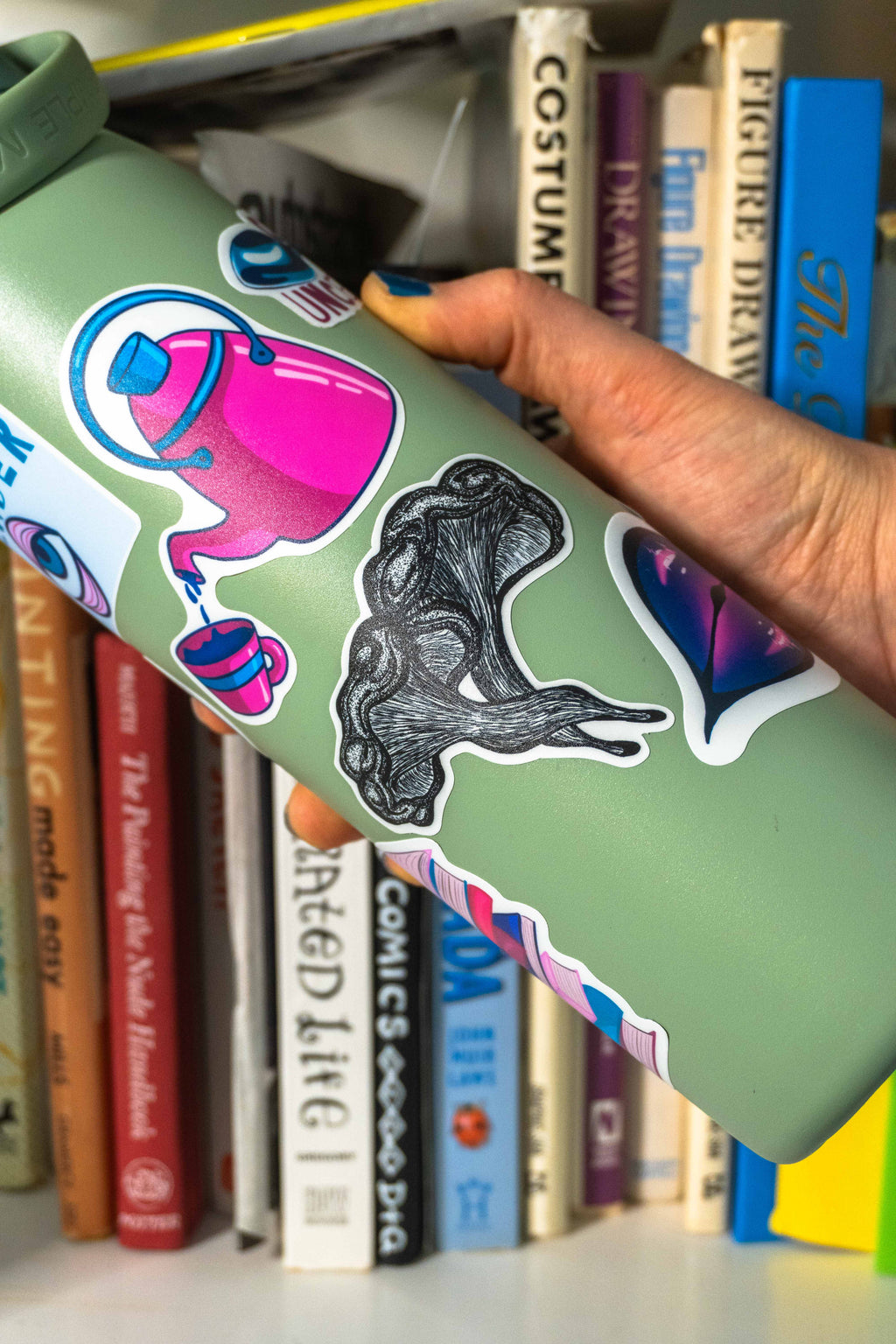 A hand with blue nail polish holds a sage green insulated water bottle decorated with colorful stickers, including a pink teapot and a grayscale mushroom sticker in the center of the bottle.