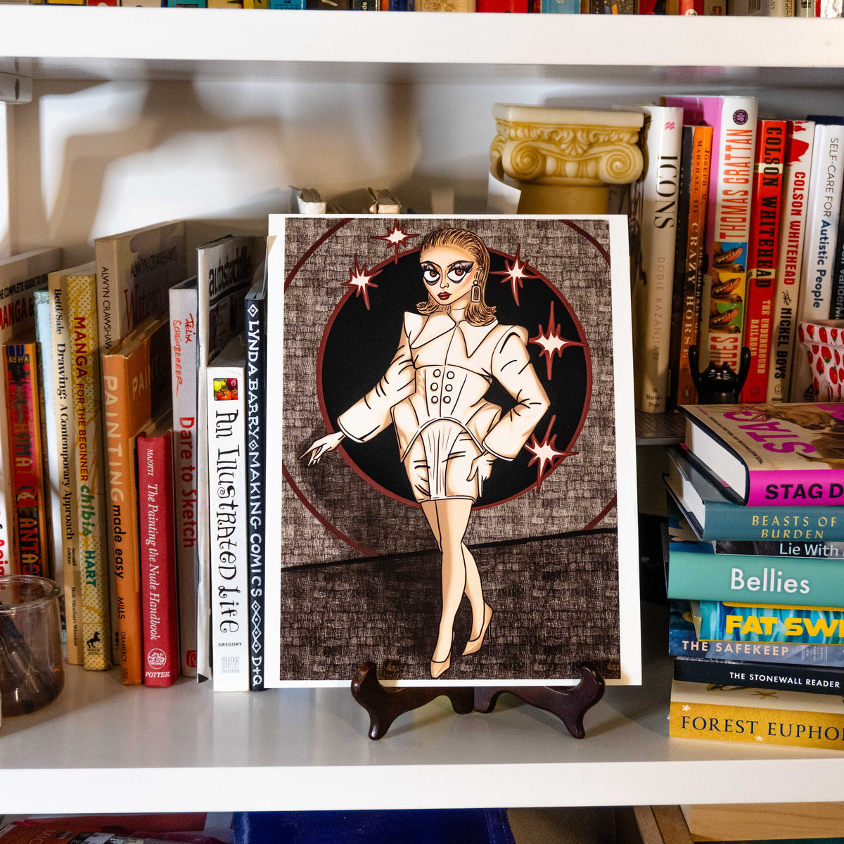 A square art print featuring a retro-style illustrated woman in a white outfit with sparkles, displayed on a small stand on a white bookshelf surrounded by books.