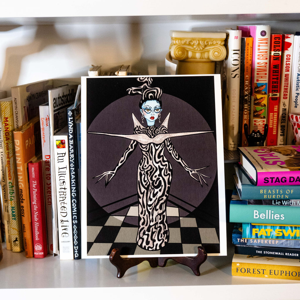 A square art print featuring a stylized black-and-white illustrated figure with dramatic blue eyes and red lips, displayed on a small stand in front of a bookshelf filled with books.