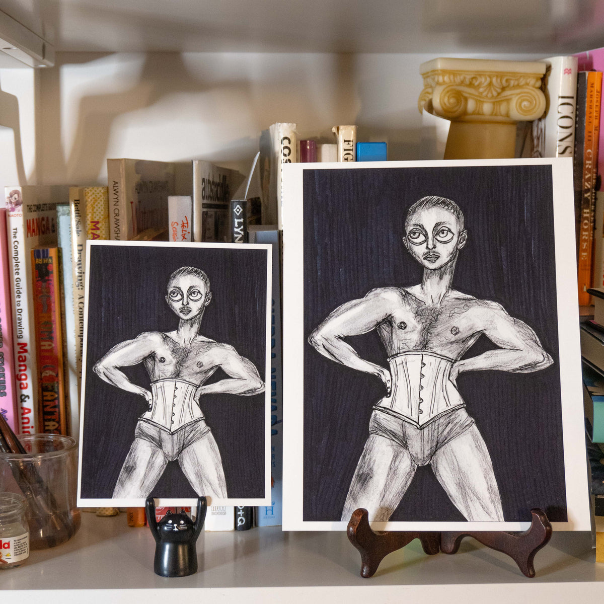 Two black and white art prints depicting a muscular figure in a corset, displayed on a shelf in front of books with small easels supporting them.