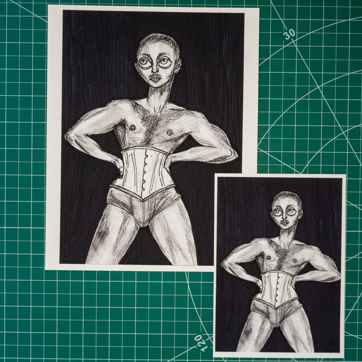 Two black and white hand-drawn sketches of a muscular figure wearing a corset and briefs, displayed on a green cutting mat background.