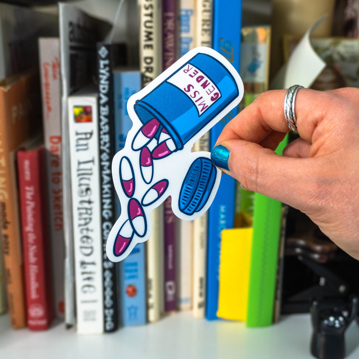 A hand with blue nail polish and silver rings holding the die cut pill bottle sticker in front of a bookshelf.