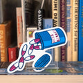 A large die-cut sticker featuring an illustrated pill bottle labelled with "Miss Gender" and large pink pills spilling out, displayed on a clear acrylic stand in front of a bookshelf.