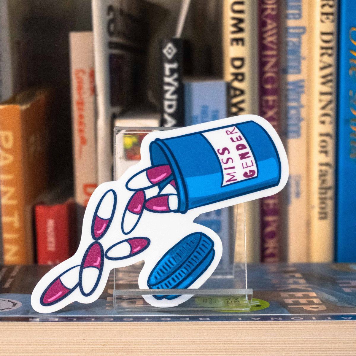 A large die-cut sticker featuring an illustrated pill bottle labelled with "Miss Gender" and large pink pills spilling out, displayed on a clear acrylic stand in front of a bookshelf.