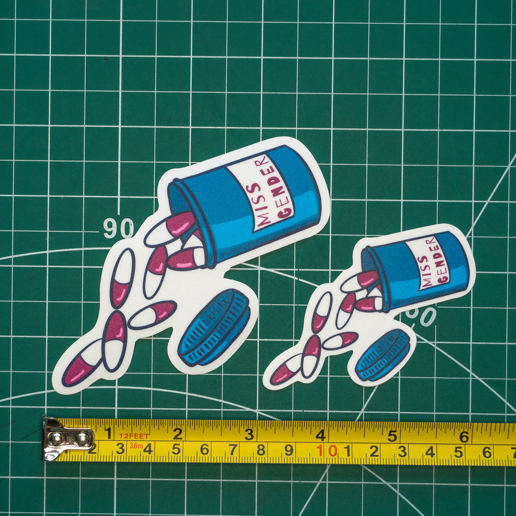 Two differently-sized die-cut stickers featuring an illustrated pill bottle labelled with "Miss Gender" and large pink pills spilling out. The stickers are placed on a green cutting mat with a yellow tape measure below for scale.