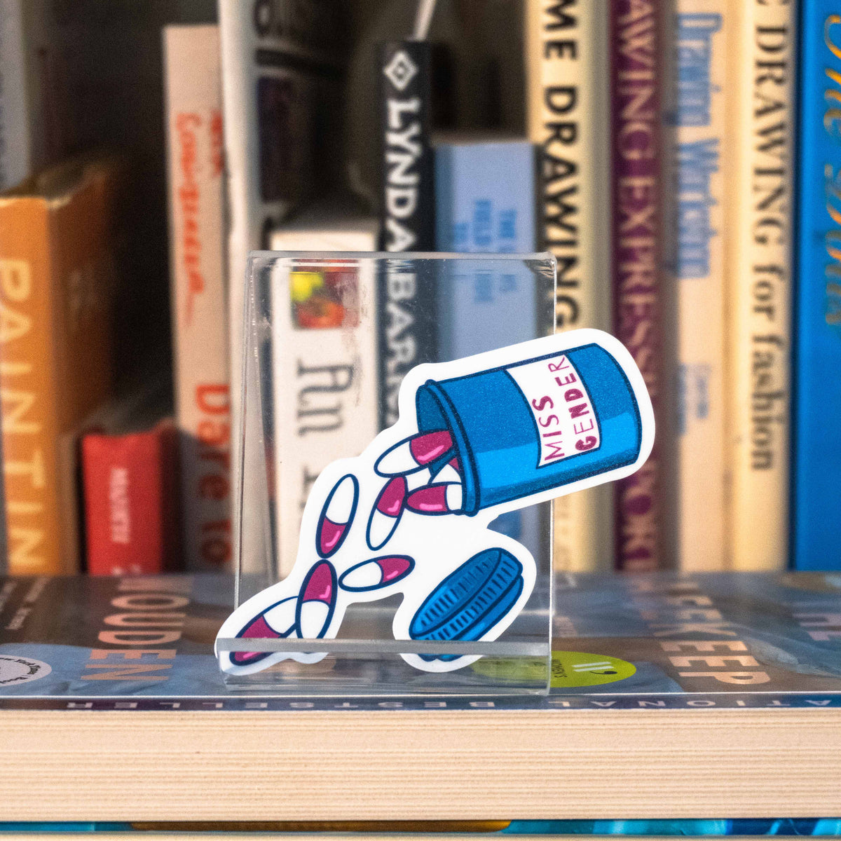 A small die-cut sticker featuring an illustrated pill bottle labelled with "Miss Gender" and large pink pills spilling out, displayed on a clear acrylic stand in front of a bookshelf.
