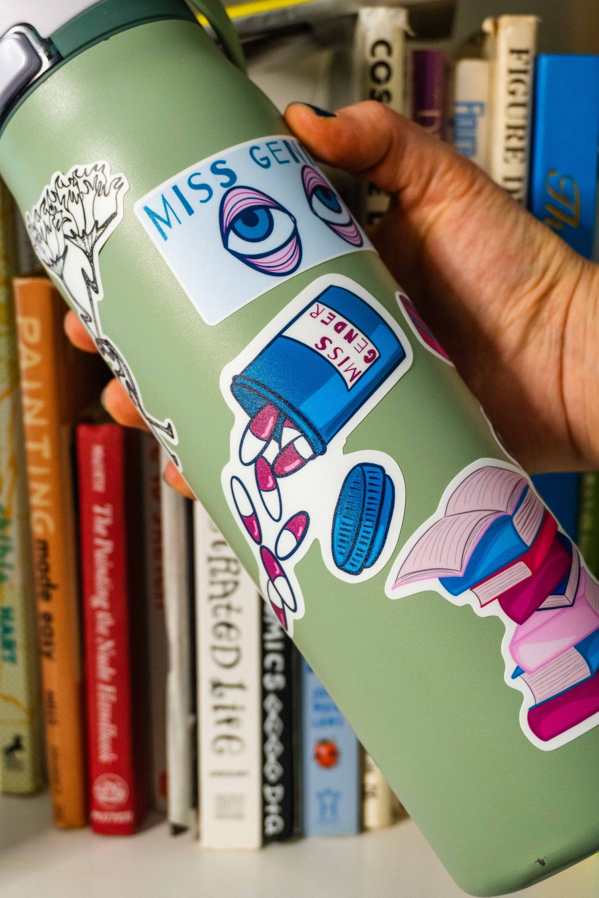 A hand holds a sage green insulated water bottle decorated with colorful stickers, withe the die-cut blue pill bottle sticker in the center of the bottle.