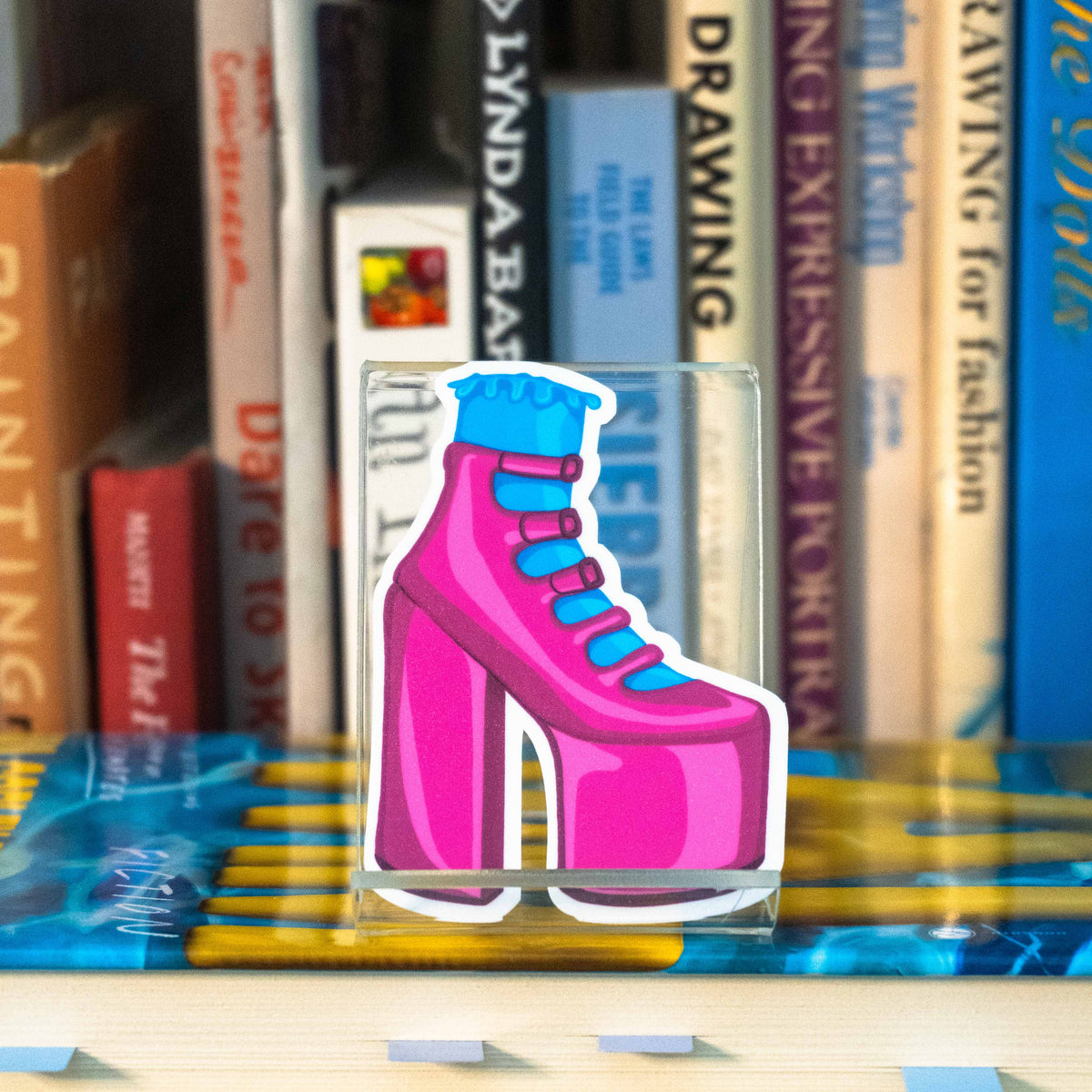 A small die-cut sticker featuring an illustrated pink strappy high heel with a blue frilly sock, displayed on a clear acrylic stand in front of a bookshelf.