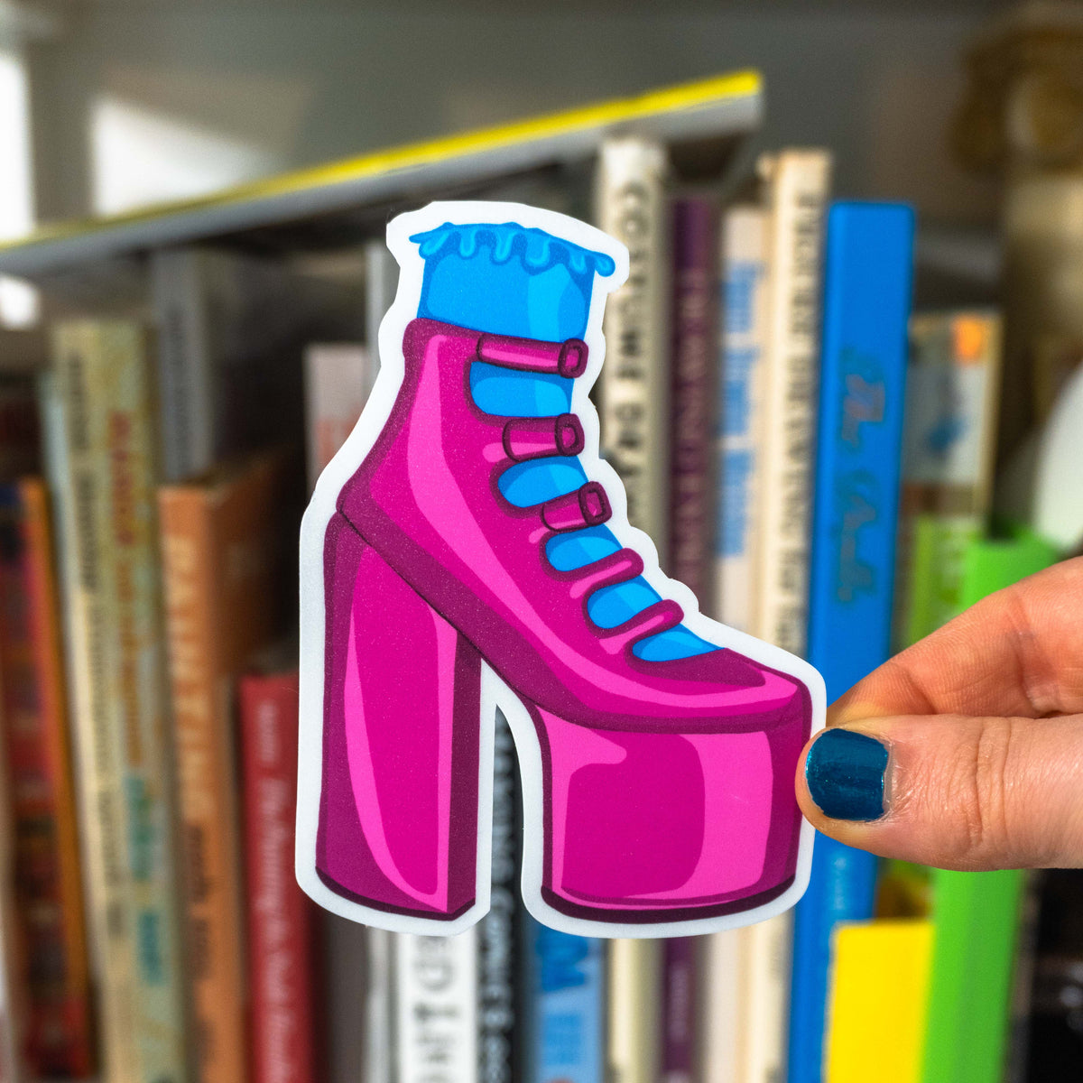 A hand with blue glitter nail polish holding the pink heel sticker in front of a background of a blurred bookshelf.