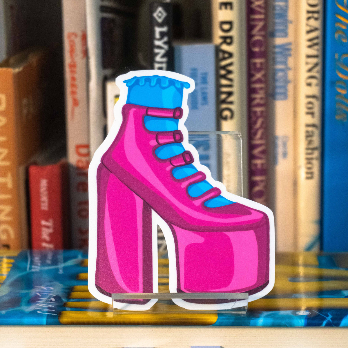 A large die-cut sticker featuring an illustrated pink strappy high heel and a blue frilly sock, displayed on a clear acrylic stand in front of a bookshelf.