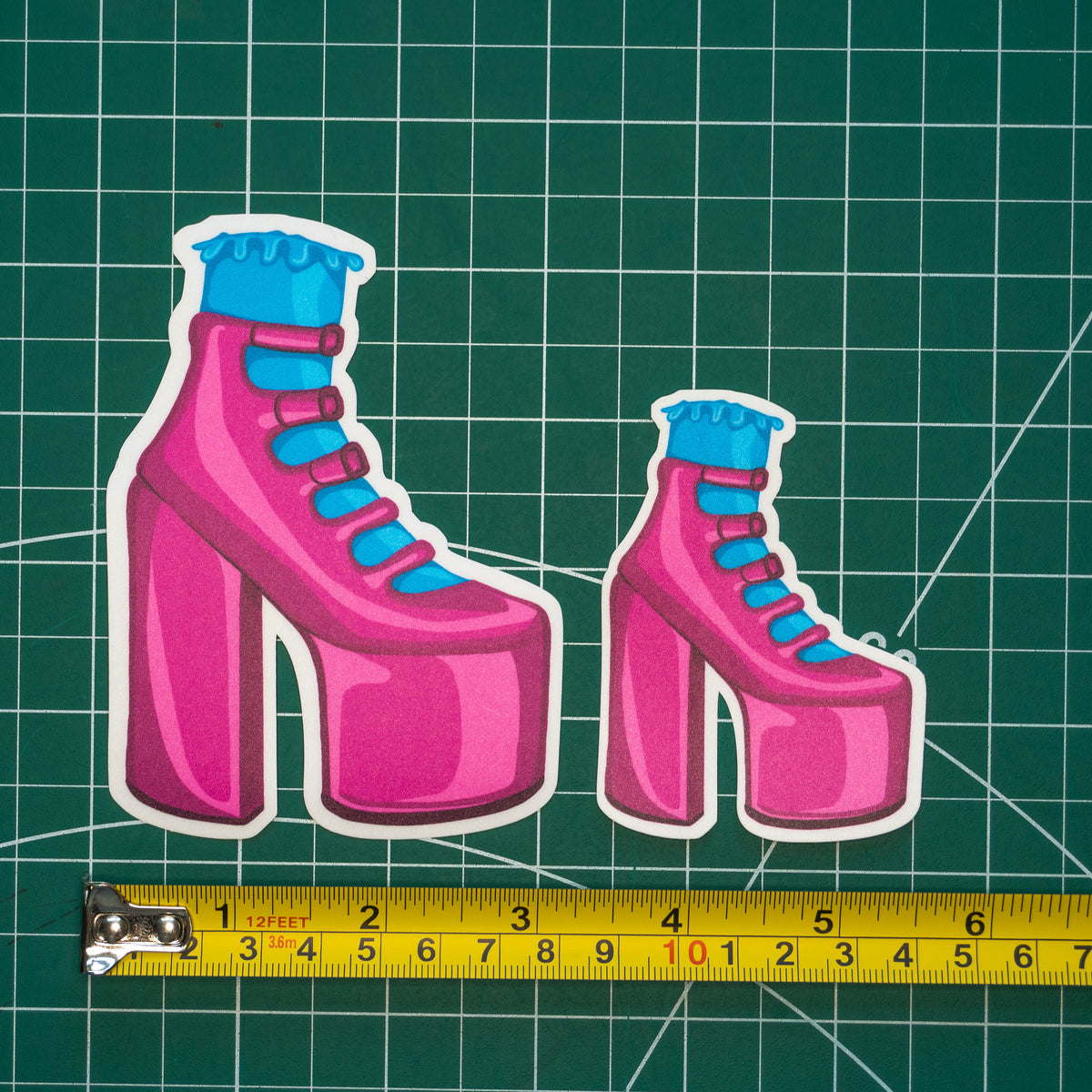 Two differently-sized die-cut stickers featuring an illustrated pink strappy high heel with a blue frilly sock placed on a green cutting mat with a yellow tape measure below for scale.