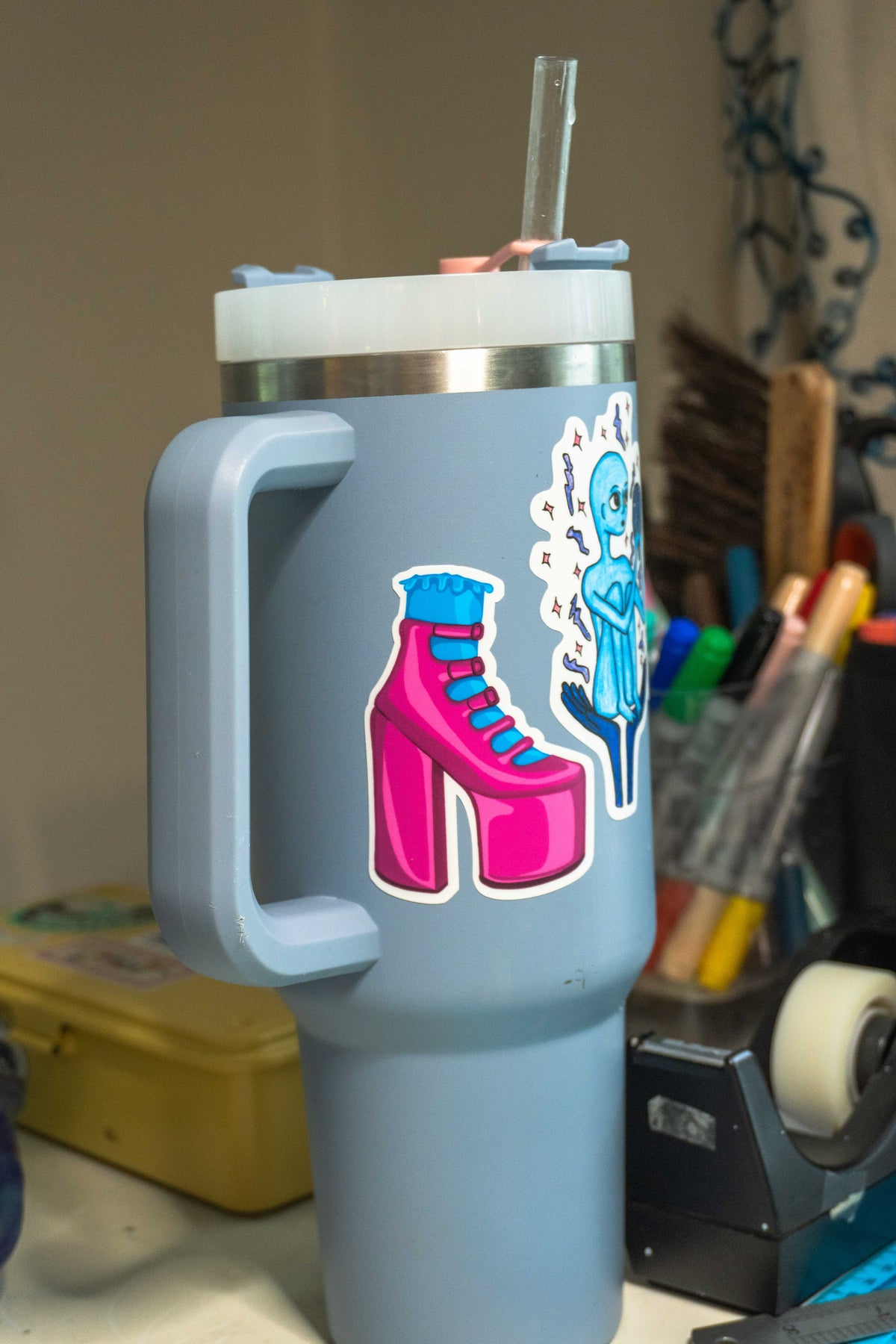 A light blue handled travel tumbler with a clear lid and straw, decorated with die-cut pink high heel sticker, set against a cluttered desk background.