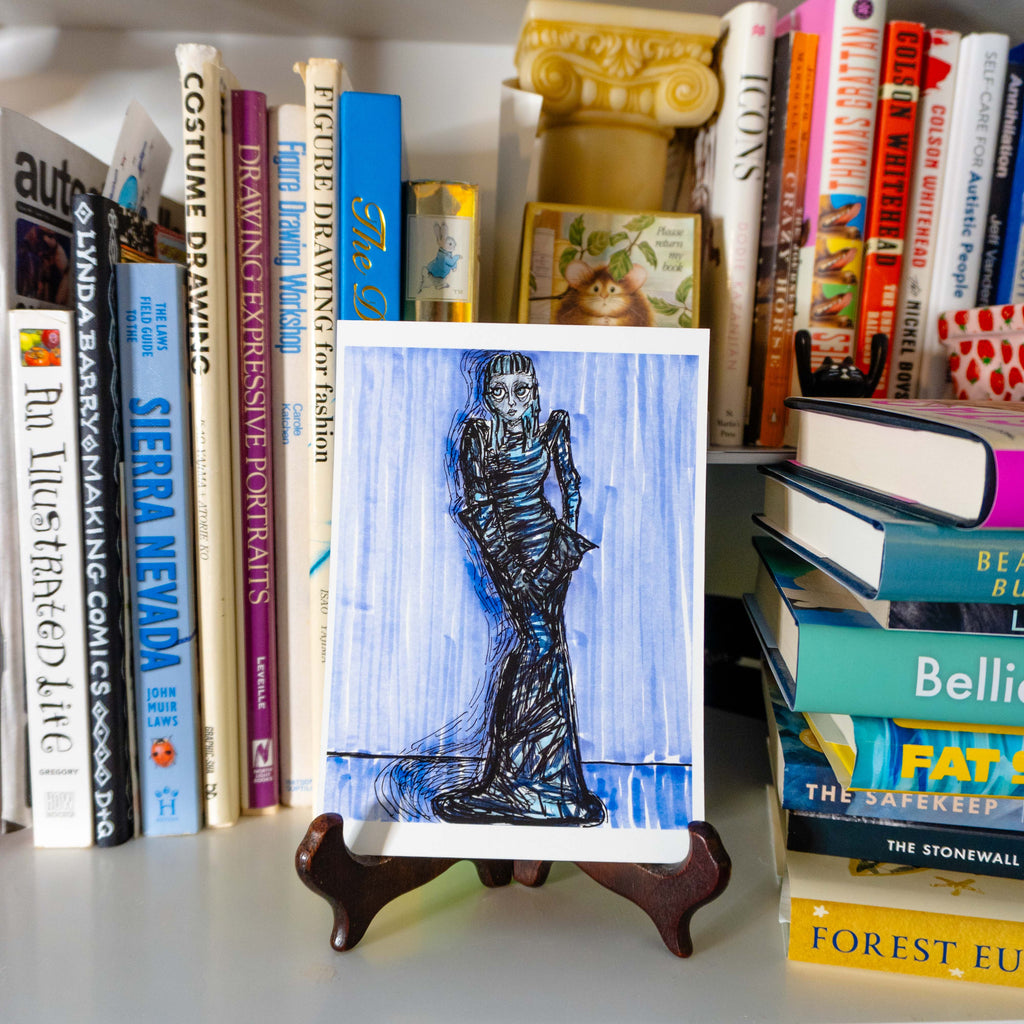 A square art print featuring a black ink sketch of a woman in a long dress on a blue watercolor background, displayed on a small wooden easel stand in front of a bookshelf.