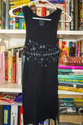 A black sleeveless tank with a black linocut print of top surgery scars and stars across the chest, hanging on a white plastic hanger in front of a bookshelf.