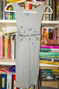 A heather gray sleeveless tank with a black linocut print of top surgery scars and stars across the chest, hanging on a white plastic hanger in front of a bookshelf.